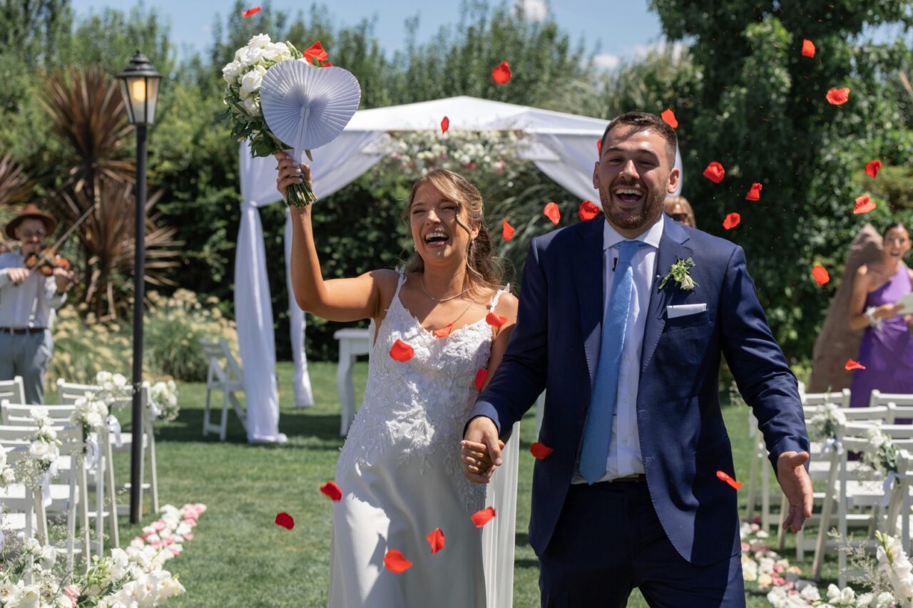 A happy couple walks down the aisle at their outdoor wedding filled with laughter and petals.
